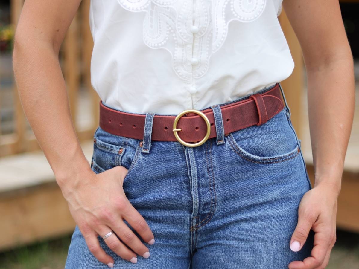 A close-up of a ladie wearing a reddish-brown leather belt with a gold-tone round buckle, styled with a white embroidered top and blue jeans.