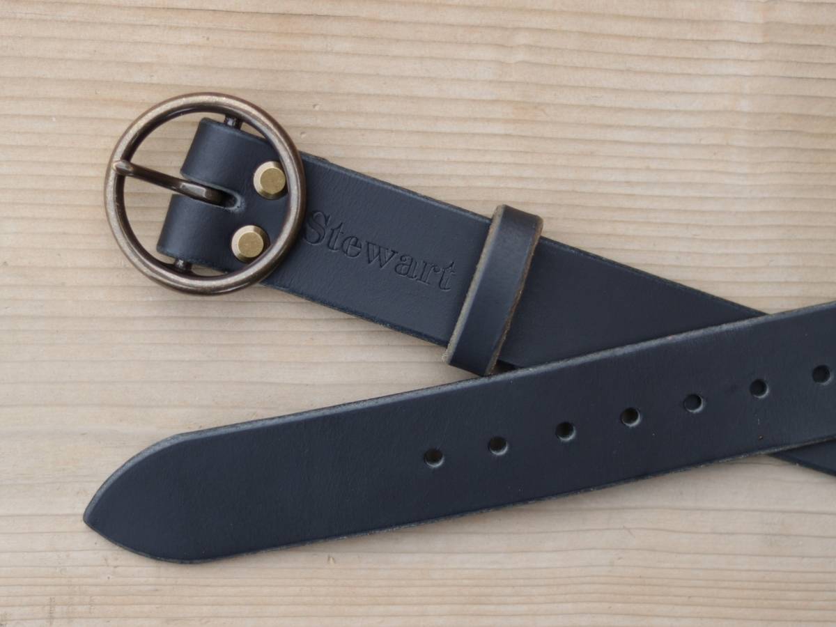 A black leather belt with a bronze-toned oval buckle and brass rivets, featuring the brand name 'Stewart' embossed on the strap, laid flat on a light wood background.