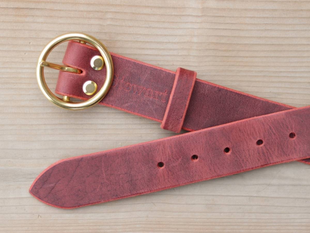 A dusty rose red leather belt with a gold-tone buckle, laid flat on a light wood grain background. The brand name 'howart' is embossed on the leather strap.