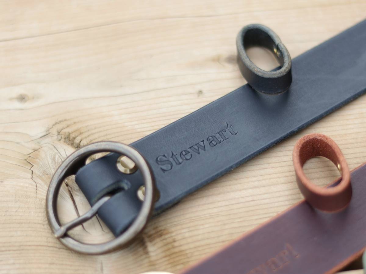 A close-up of a black leather belt with a round metal buckle and the name 'Stewart' engraved on the strap, placed on a light wood surface.