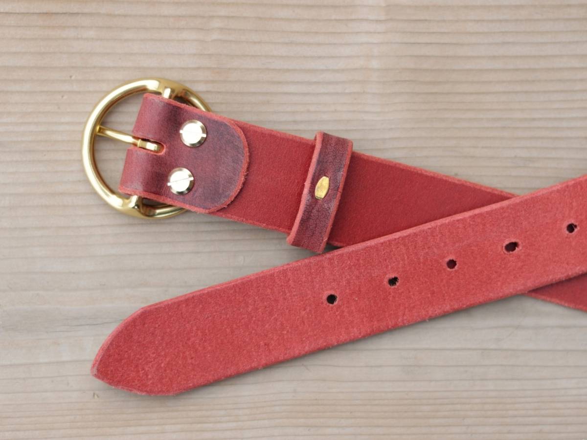 A red suede belt with a gold-tone metal buckle and silver-toned rivets, laid flat on a light wood background.