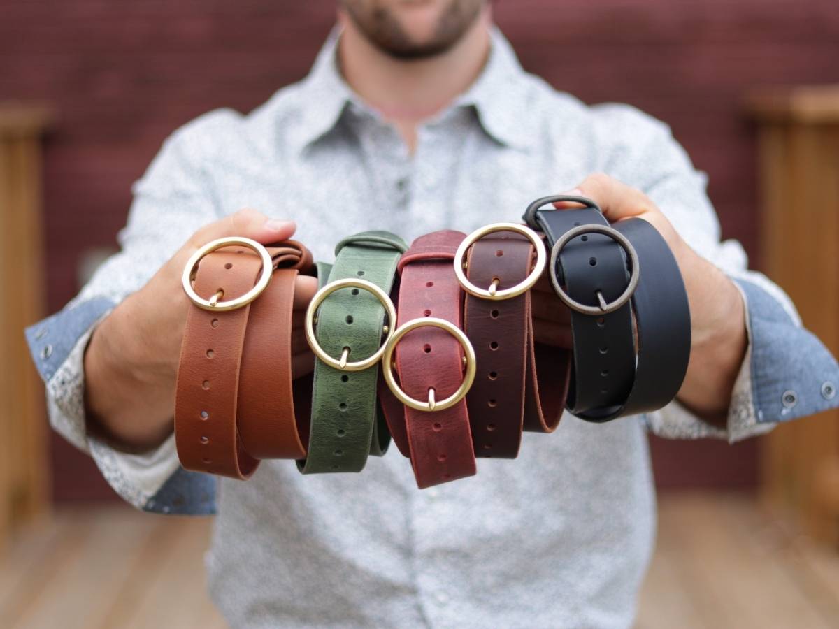 Charles from Stewart Leather holding all five Her Belt colours — Espresso, Black, Cognac, Sage Trail, and Vintage Oxblood water buffalo leather belts with brass buckles