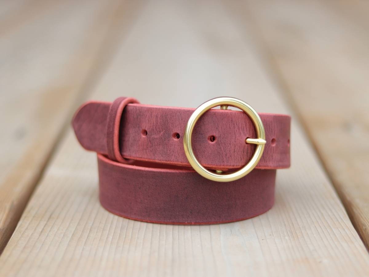 A dusty rose ladies leather belt with a gold-tone round buckle, coiled on a light-colored wooden surface.