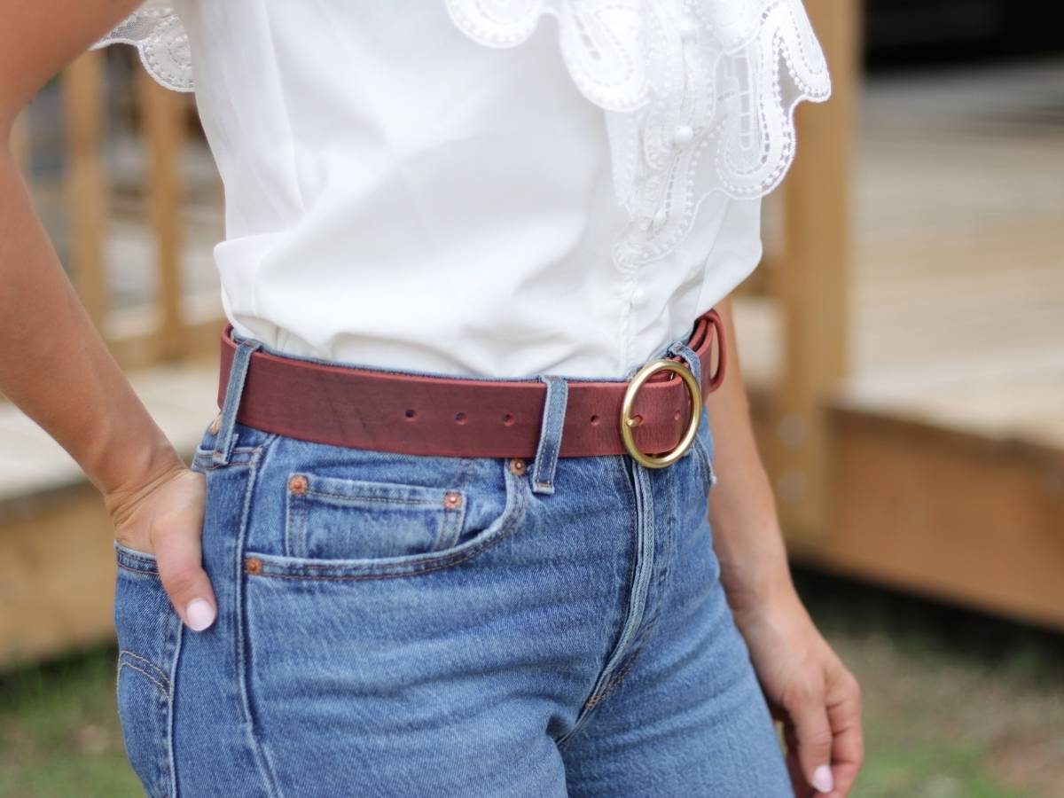 A ladie wearing a white blouse with lace trim, blue jeans, and a brown leather belt with a gold-tone round buckle, standing outdoors near a wooden structure.