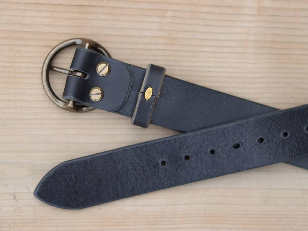 A close-up of a dark navy leather watch strap with a round bronze buckle and gold-tone hardware, laid diagonally on a light wood grain background.