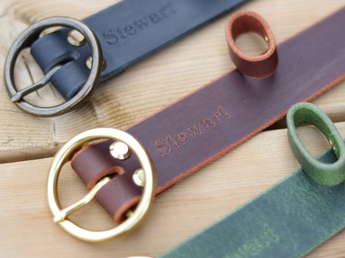 Close-up of brown leather belt laid diagonally on a light wood surface, each with a different buckle color and a small matching loop detail. The belts are dark blue, brown, and green, and the name 'Stewart' is embossed on the leather.