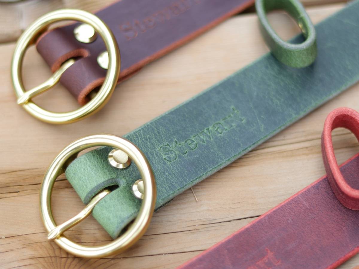 Three leather belts with brass buckles arranged diagonally on a wooden surface. The belts are brown, green, and red, with the green belt in the foreground showing the brand name 'Stewart' embossed on the strap.