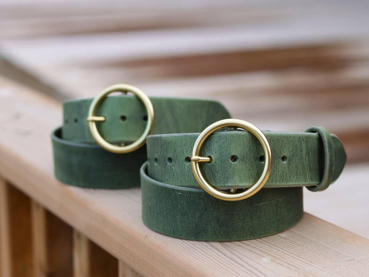 Two dark green ladies leather belts with large gold-tone round buckles, coiled on a light wooden surface with a softly blurred background.