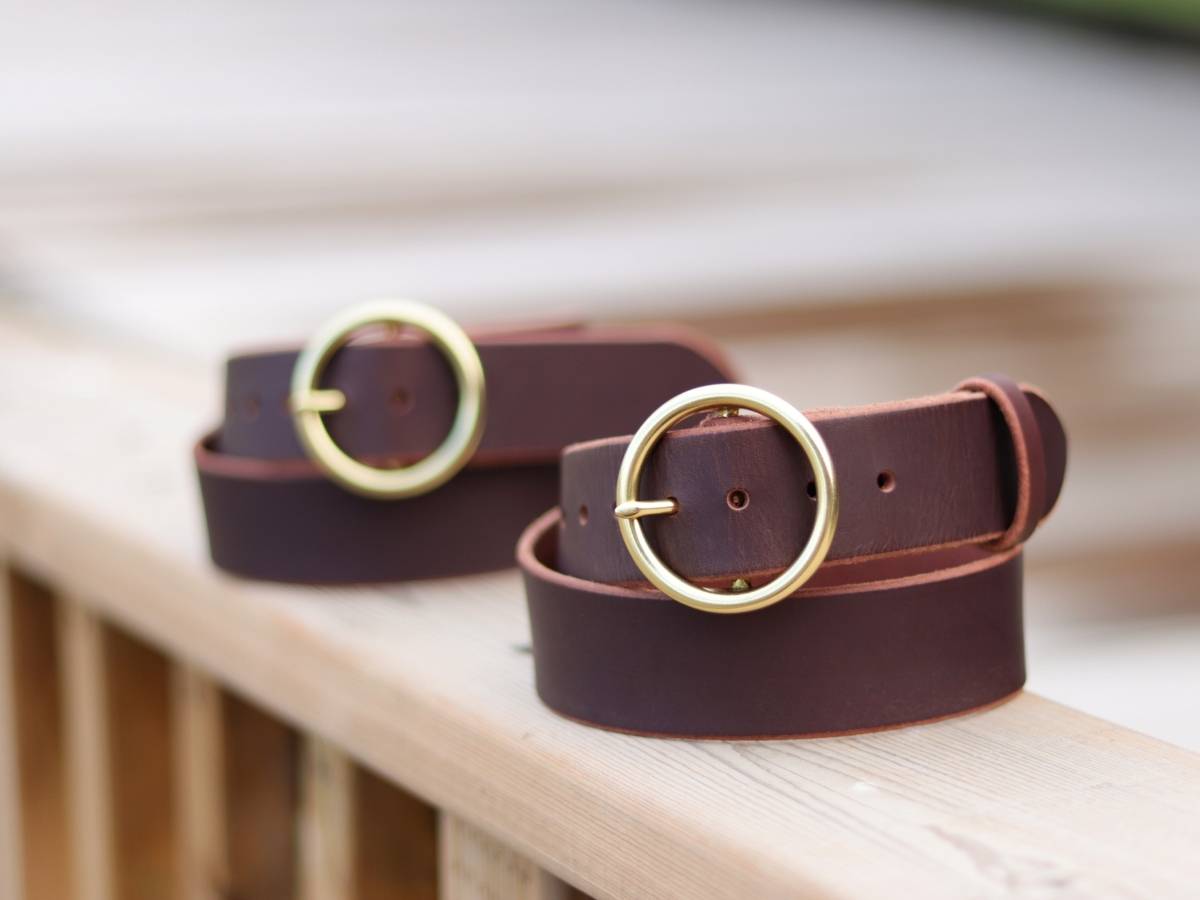 Two dark brown ladies leather belts with round gold-tone metal buckles resting on a light wooden surface.