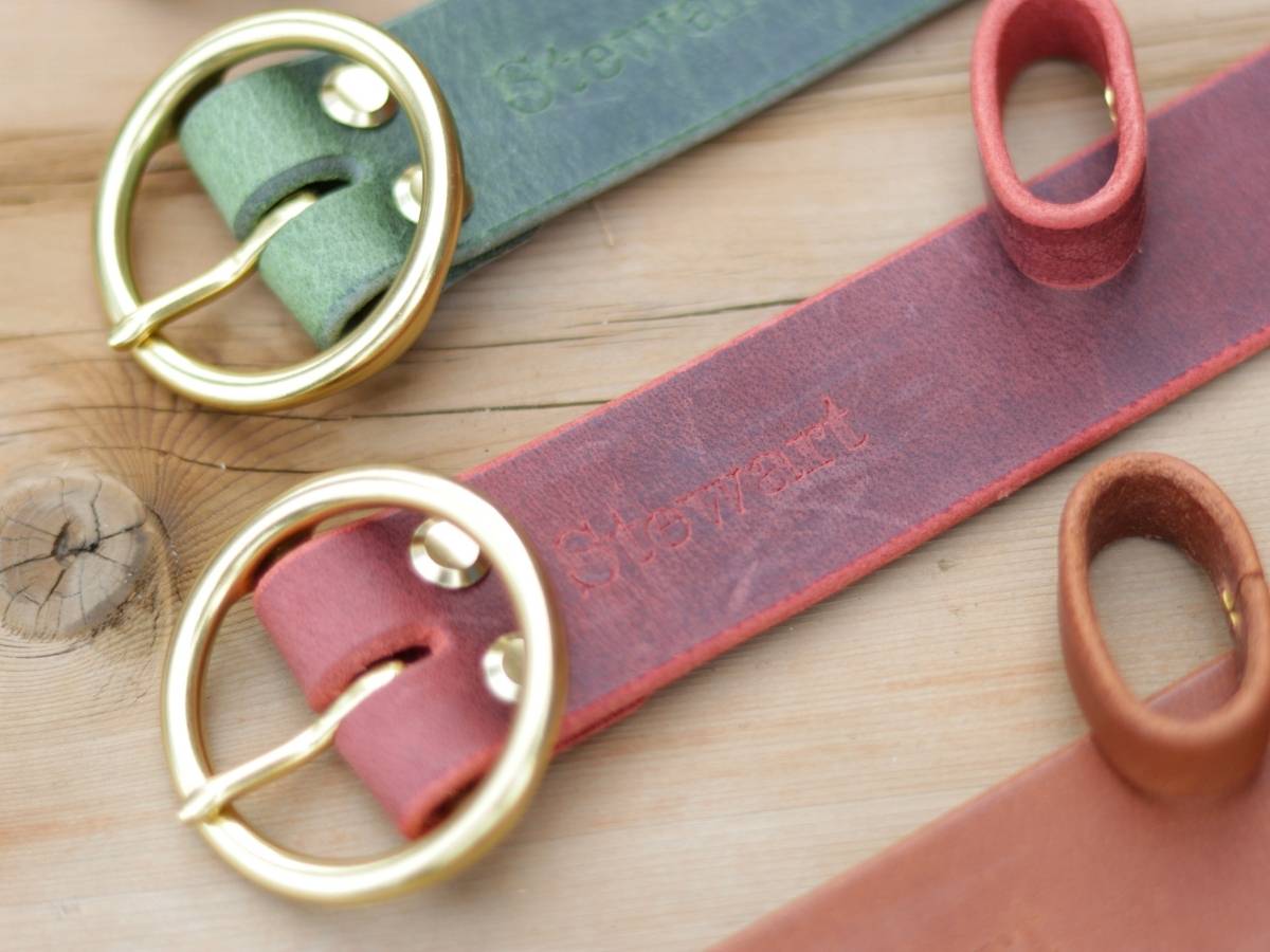 Close-up of ladies leather belts with brass buckles laid on a wooden surface. Visible embossed brand text reads 'Stewart'.